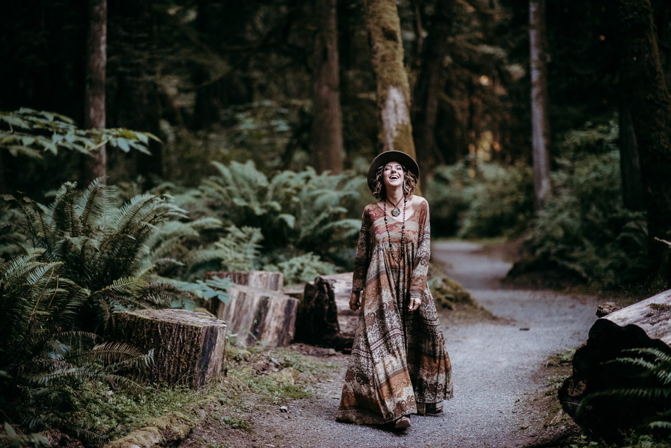 woman with curly brown shoulder length hair wearing floor length boho dress and a wide brimmed hat walks through old growth forest with personal brand photographer sherry nelsen