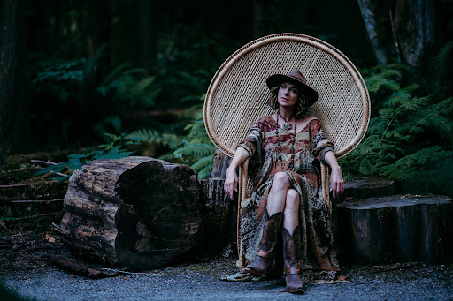 allana pratt,  wearing a long dress and cowboy boots sitting on a wicker chair in the forest during a personal branding session with sherry nelsen photography.