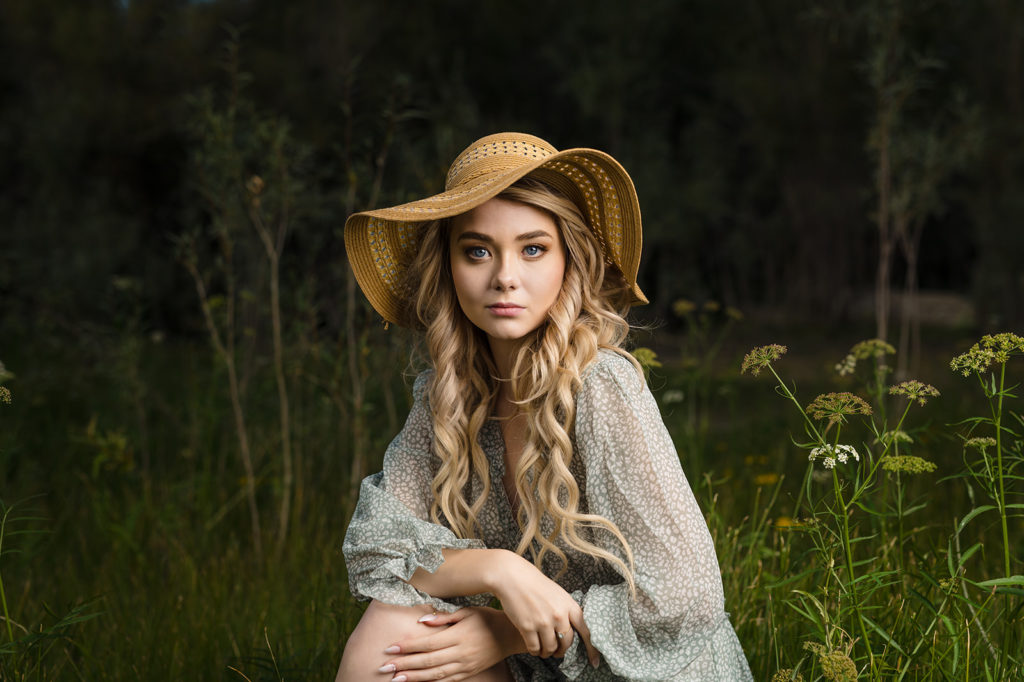 young white woman with long flowing blonde hair wearing a flowing wide brimmed mustard yellow hat and a flowing dress crouches in a dill patch
